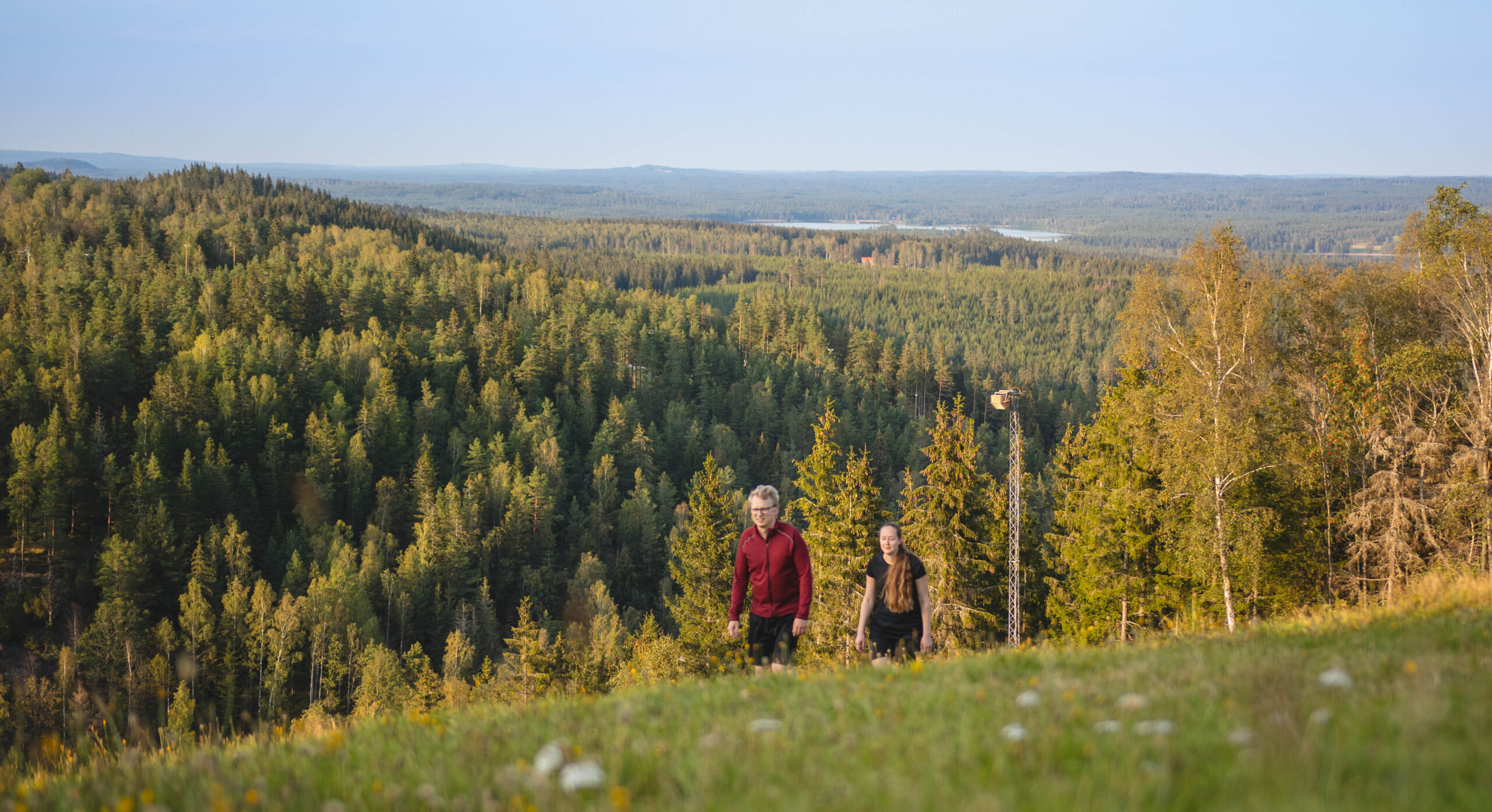 Två personer på väg upp mot toppen på Isaberg med majestätisk skog bakom sig och med Algustorpasjön skymtandes en bit bort