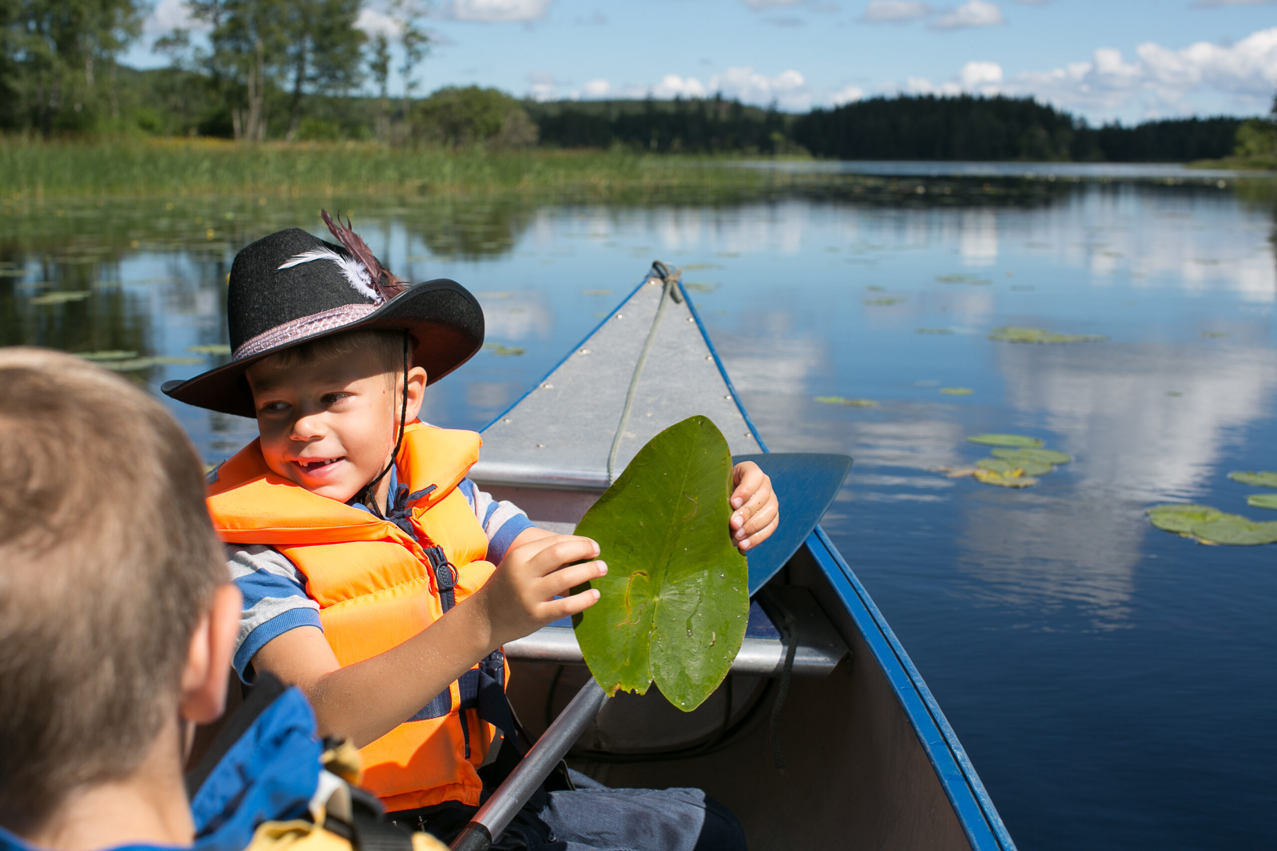 Två pojkar på kanot vid Hestraviken hotell i Småland, där en pojke håller upp ett näckrosblad efter en spännande tur på vattnet.