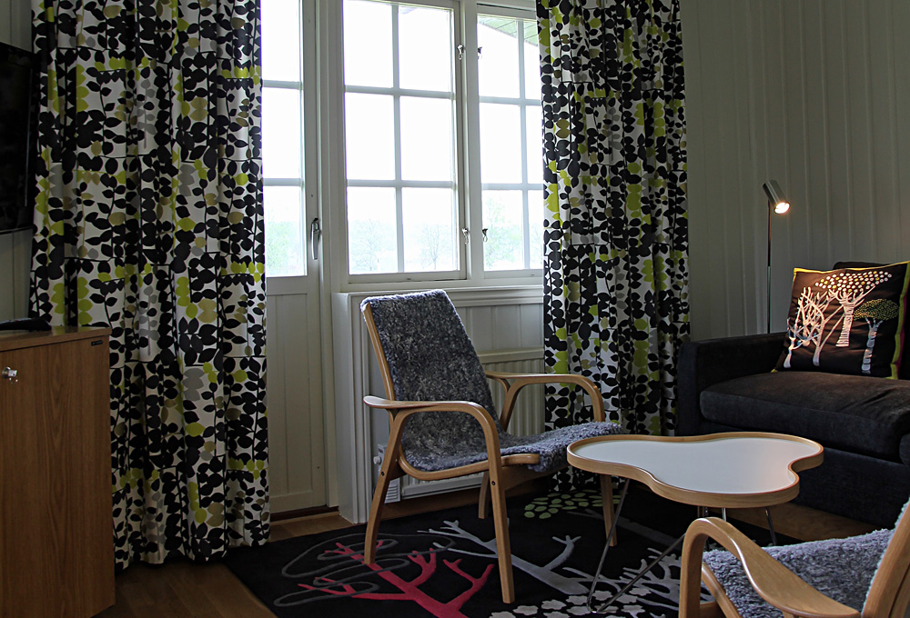 Salongen i våra Fristående Stugor med bäddsoffa, laminofåtöljer, katheamatta och med altandör som leder ut till terassen mot trädgåden och Algutsorpa sjön