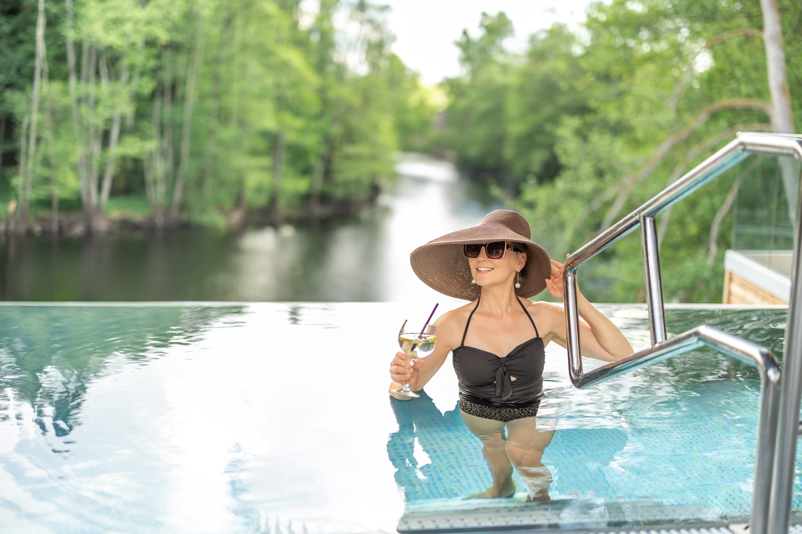 Kvinna i solhatt med drink i handen i infinitypoolen på Hestraviken, med Nissan i bakgrunden och en avkopplande atmosfär.