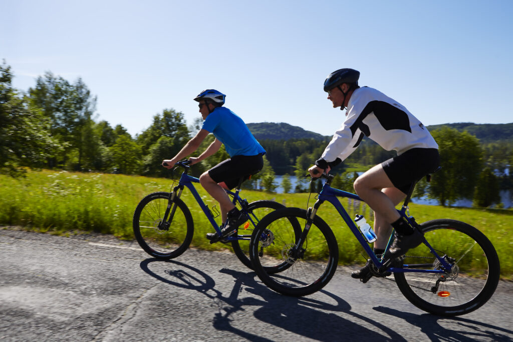 Två mtb cyklister en solig dag utmed Pink Lady där en del av leden är landsväg, Algustorpasjön och Isaberg i bakgrunden