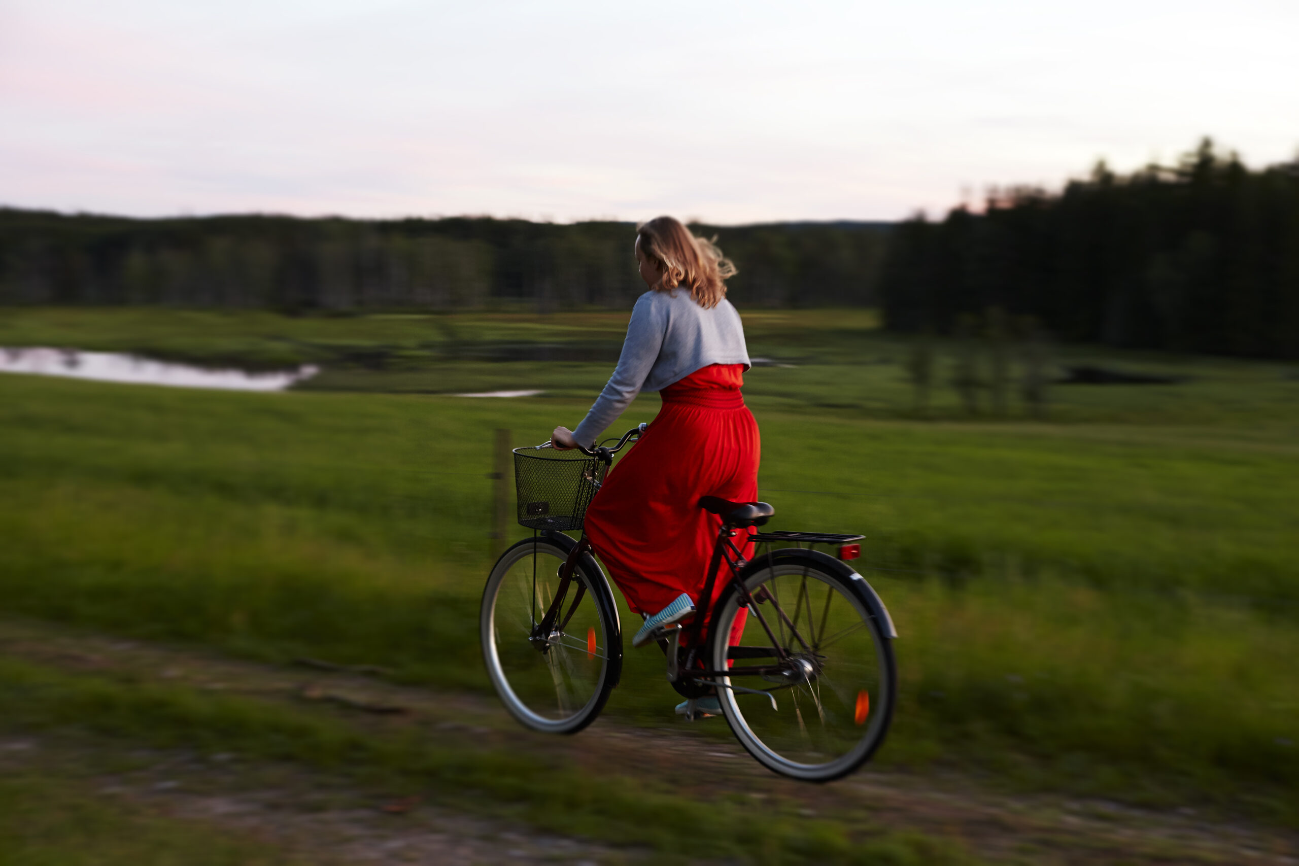 Kvinna på cykel på grusväg i röd klänning en sommarkväll, cykel lånad från Hestraviken