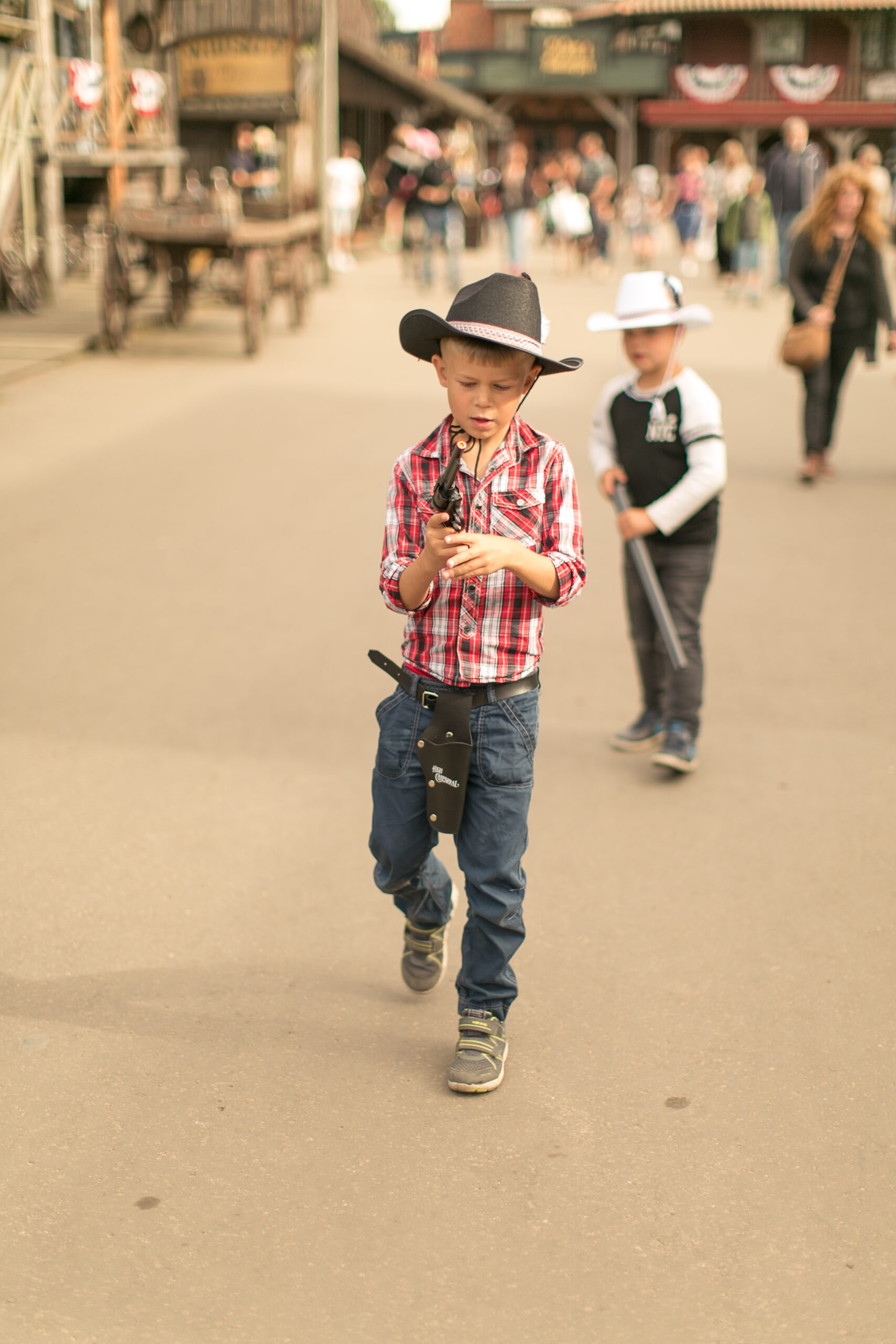 Barn i rutig skjorta med cowboyhatt, hölster och leksakspistol som promenerar på huvudgatan, fler barn och vuxna syns i bakgrunden i Western staden High Chaparall