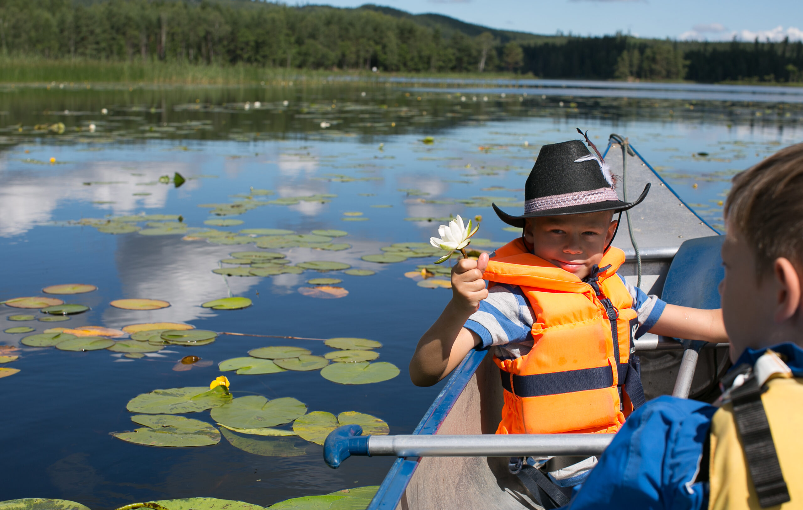 Två barn med flytvästar på sig paddlandes i kanot på Algustorpasjön, ett av barnen håller upp en näckrosblomma