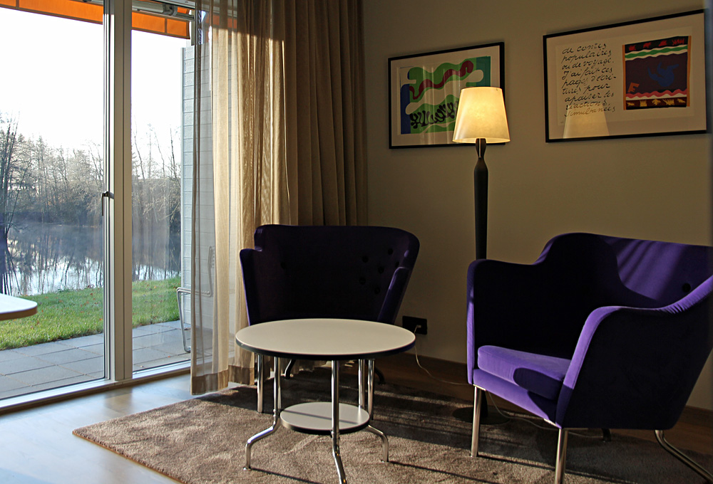 Dubbelsäng med panoramafönster, sittfåtöljer samt terass med möbler från Grythyttan och utsikt över Nissan i våra Dubbelrum Riverside i Villa 5