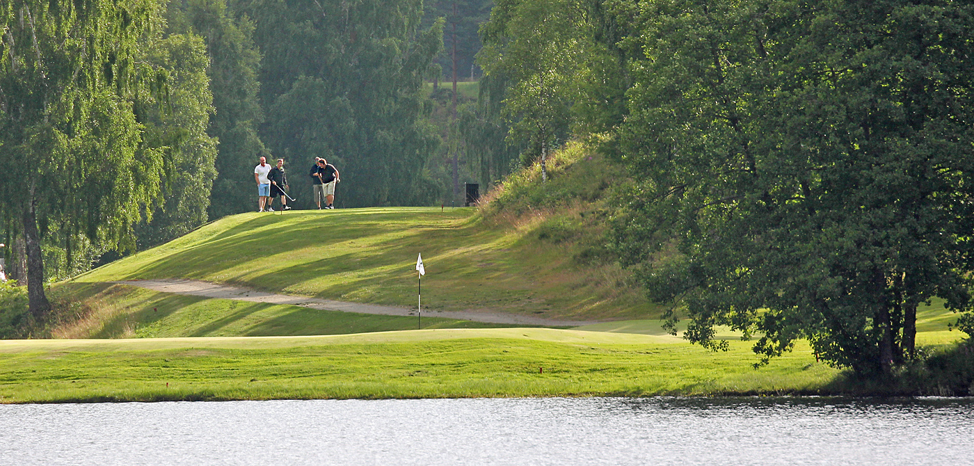 Tre golfspelare spelar mot greenen vid Hammarsjön på Isabergs golfbana en solig sommardag, omgiven av Smålands vackra natur.