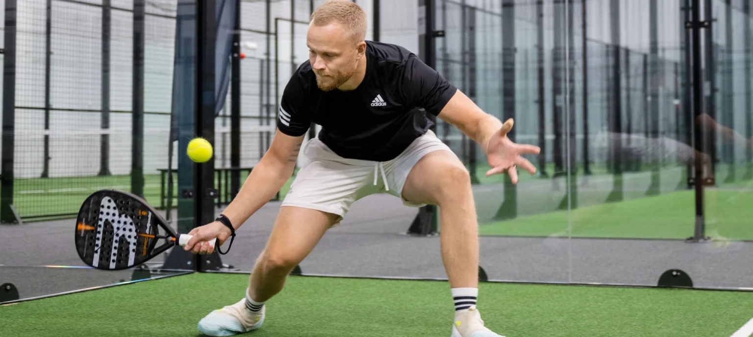 Man slår ett backhand slag i en pågående paddelmatch på en inomhusbana i närheten av Hestraviken, flera banor syns i bakgrunden